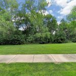 A serene outdoor scene featuring a grassy area with a sidewalk, surrounded by lush green trees and a partly cloudy sky.