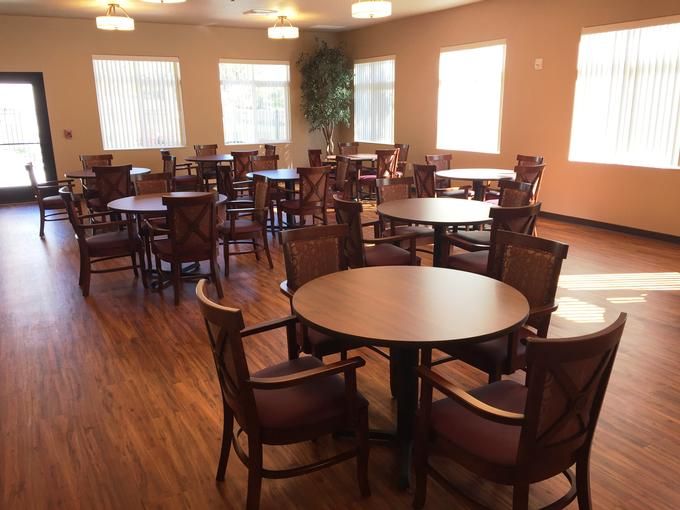 An empty dining room with round tables and wooden chairs, lit by natural light through multiple windows.
