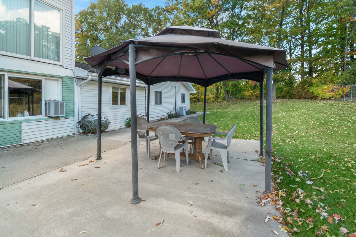 An outdoor patio area with a gazebo covering a wooden table and chairs set, surrounded by a grassy lawn.