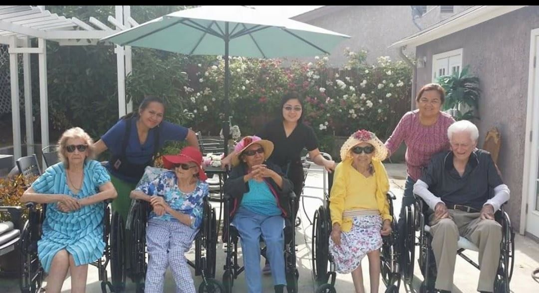 A group of elderly individuals in wheelchairs accompanied by caregivers, enjoying time outdoors under a large umbrella.