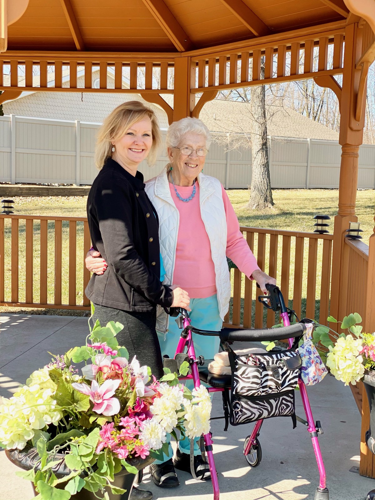 An elderly woman and a young woman standing together outdoors in a gazebo surrounded by flowers, with the elderly woman using a walker.