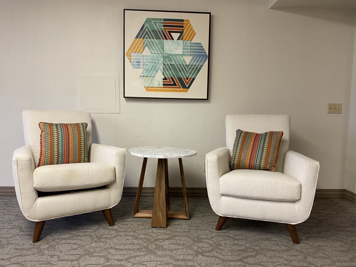 A modern interior setting with two white armchairs, a small wooden table, and geometric artwork on the wall.
