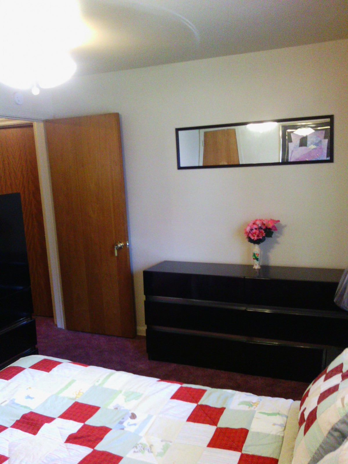 A cozy bedroom featuring a dresser with a vase of flowers, a bed, and a mirror on the wall.