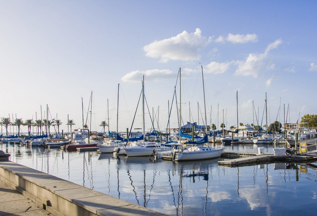 A tranquil marina with sailboats reflected on calm water under a partly cloudy sky.