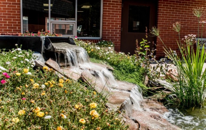 A small waterfall cascading through a garden with flowers in front of a brick building.