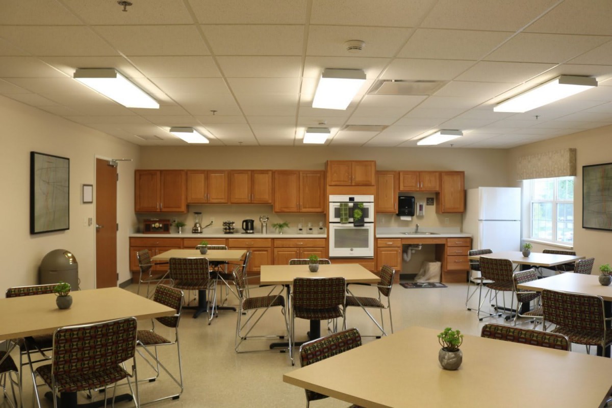 A community kitchen area with tables and chairs, featuring wooden cabinets and overhead lighting.