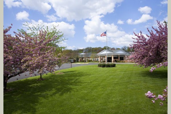 A scenic view of a building with an American flag, surrounded by blooming trees and a green lawn under a cloudy sky.