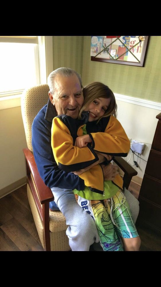 An elderly man and a child share a warm embrace while sitting in a chair indoors.