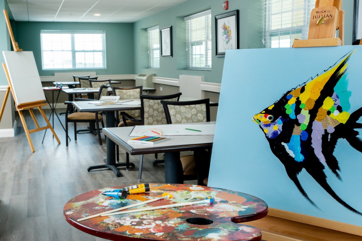 An art classroom with tables and chairs, featuring a painting on an easel and various art supplies.