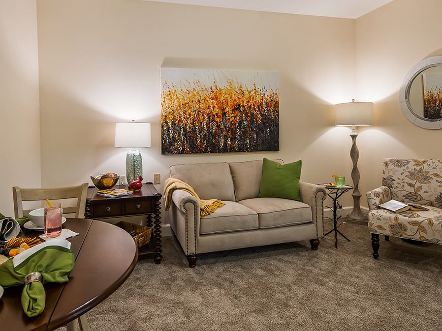 A cozy living room with a beige sofa, decorated with green pillows, flanked by lamps and a vibrant nature painting on the wall.