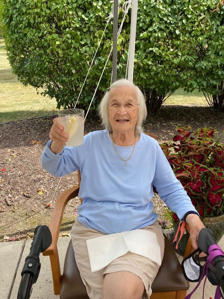 An elderly woman sitting outdoors, smiling and holding up a drink.