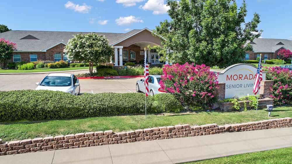 A scenic view of a building with a parking lot, surrounded by trees and landscaping, featuring a flagpole and flowering bushes.