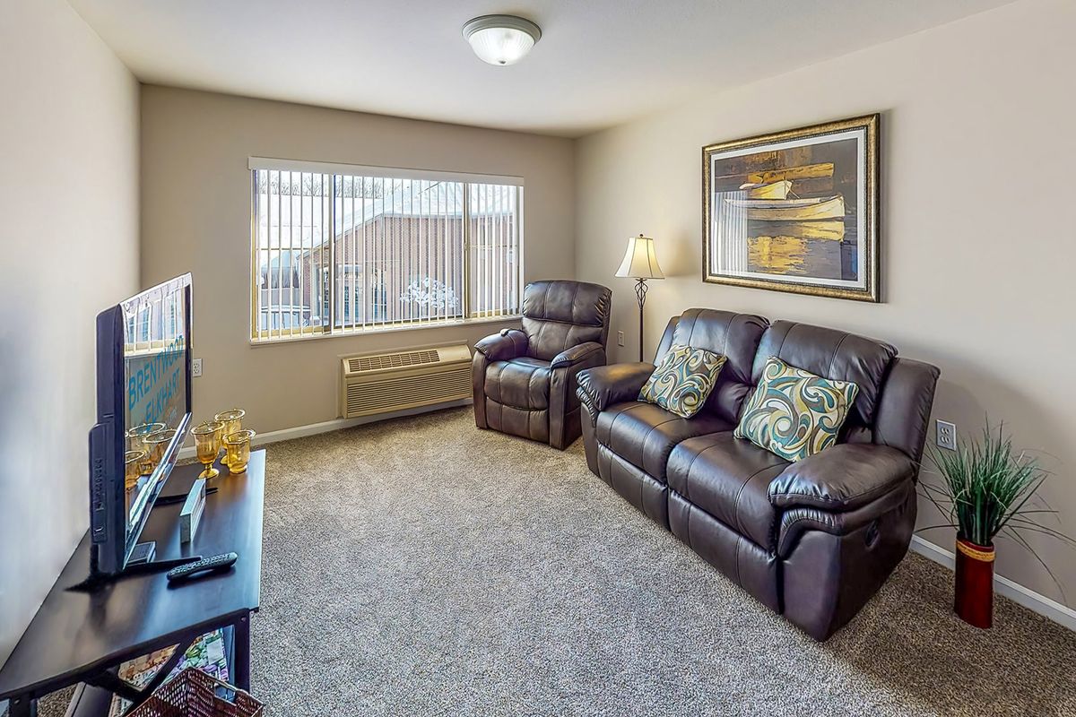 A cozy living room with a sofa, armchair, and a TV, featuring large windows and a decorative painting on the wall.