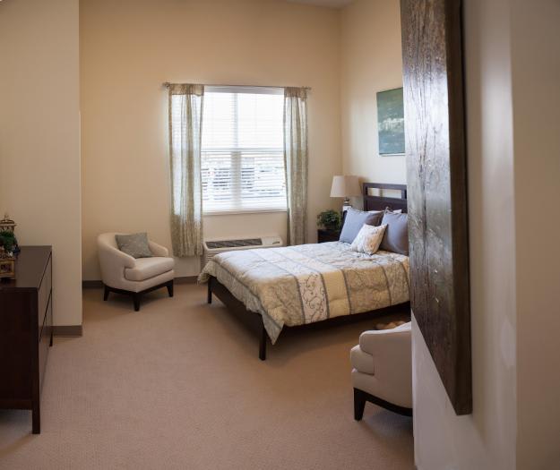 A cozy bedroom with a neatly made bed, two chairs, and a window with sheer curtains.