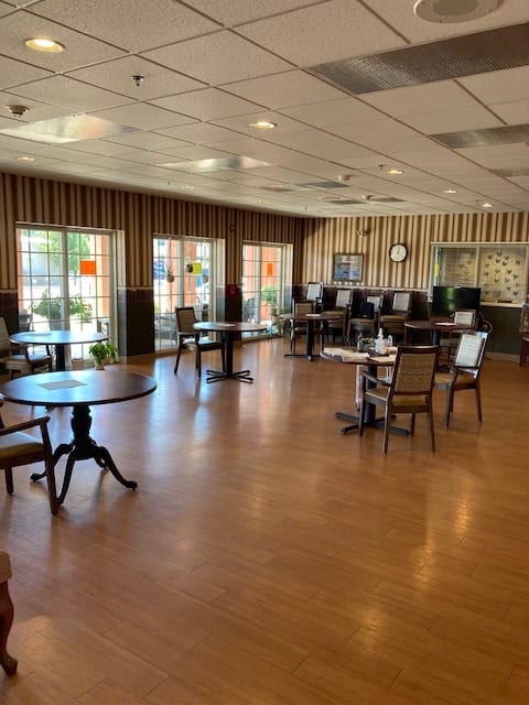 A neatly arranged dining area with tables and chairs on a wooden floor, featuring decorated walls and large windows.
