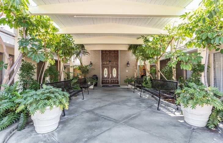 A covered entrance with benches and lush greenery leading to a set of wooden doors.