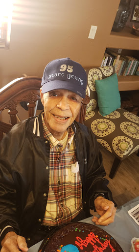 An elderly man wearing a blue cap and a plaid shirt, smiling while seated indoors.