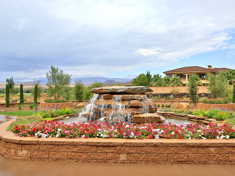 A beautifully landscaped garden with a stone fountain and vibrant flowers, set against the backdrop of a large building and cloudy sky.