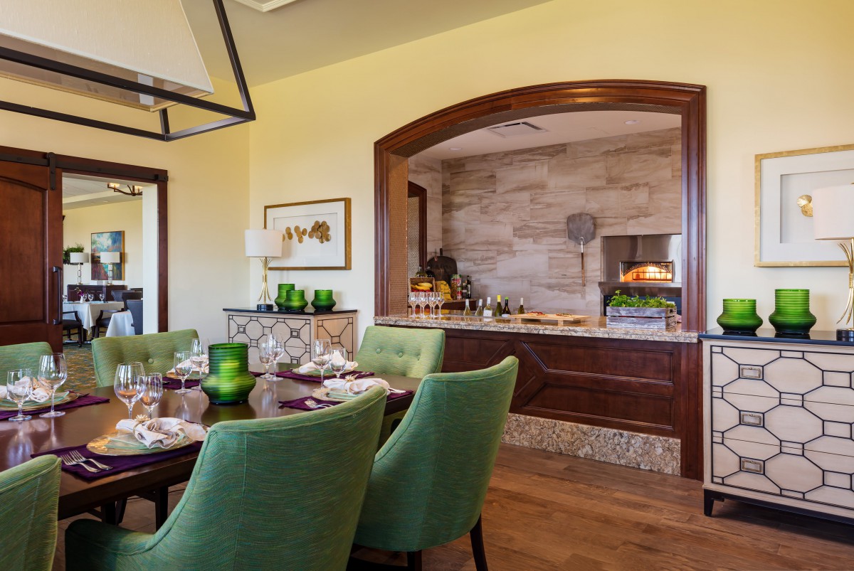 A modern dining room with green chairs and a well-set table, featuring a kitchen alcove with a pizza oven.
