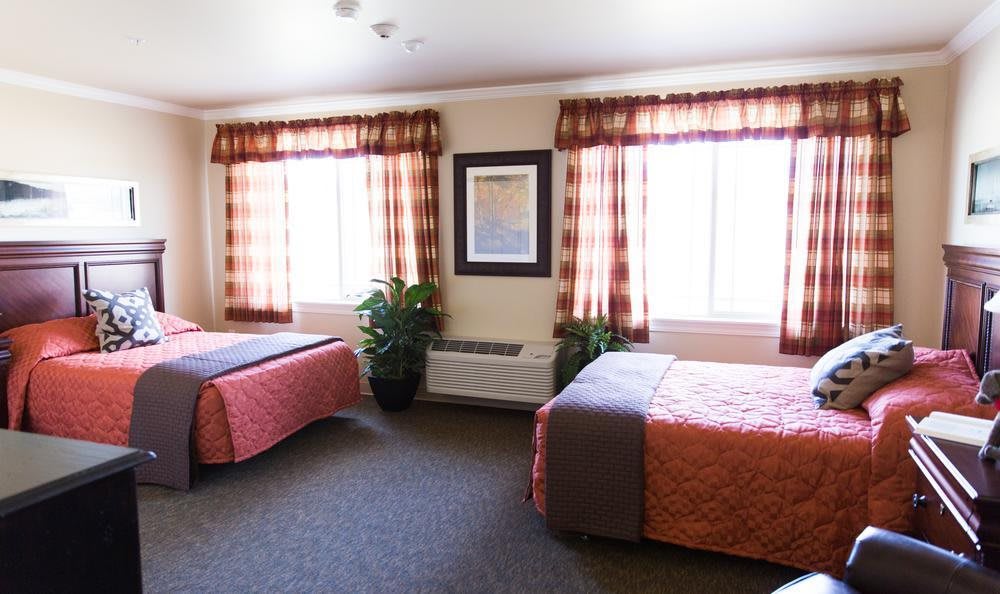 A bright hotel room featuring two neatly made beds with patterned bedding, large windows with curtains, a framed picture on the wall, and a carpeted floor.