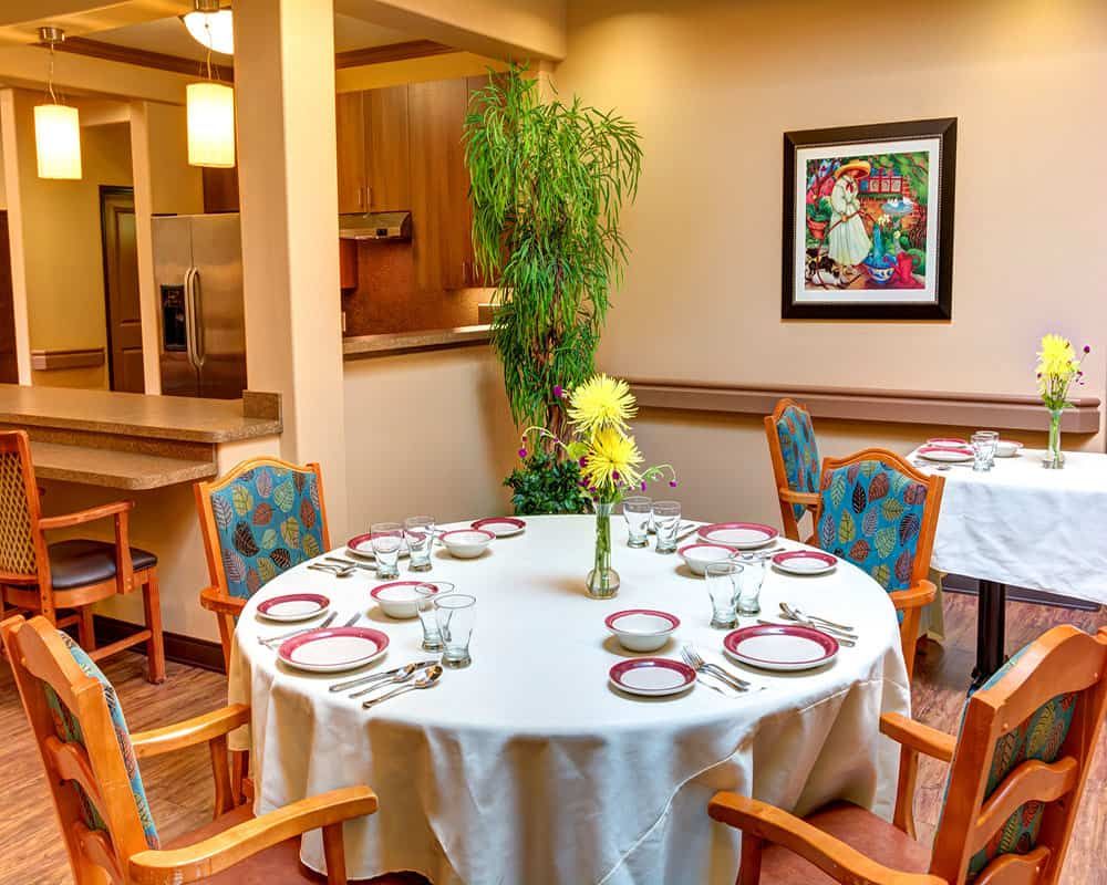 A warmly decorated dining room with a round table set for a meal, featuring elegant tableware, wooden chairs, and a vibrant painting on the wall.
