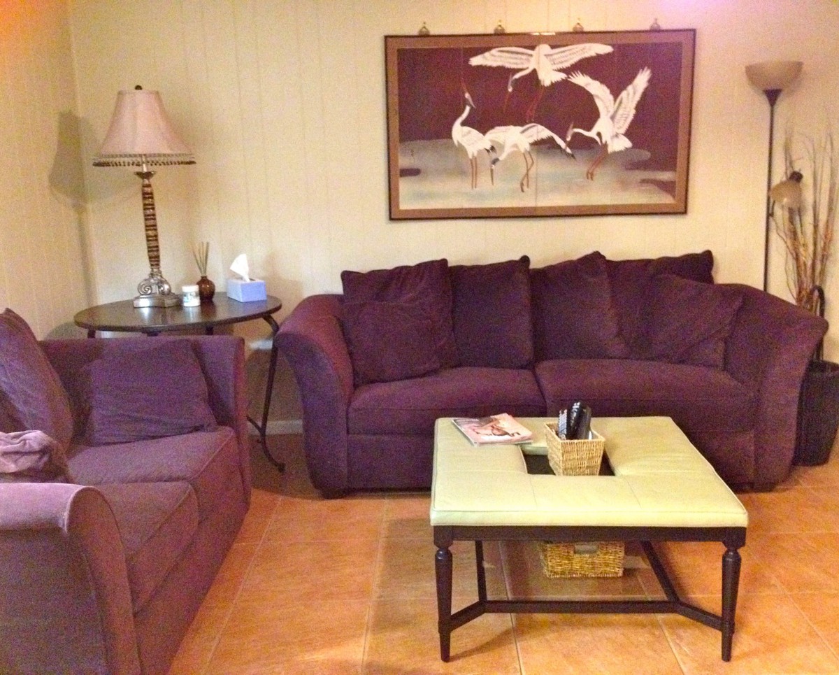 A cozy living room with a purple sofa, matching armchair, and a coffee table, featuring wall art and a floor lamp.