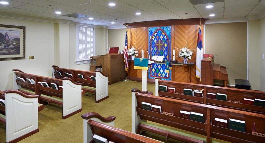 A small chapel interior with wooden pews and a stained glass backdrop.
