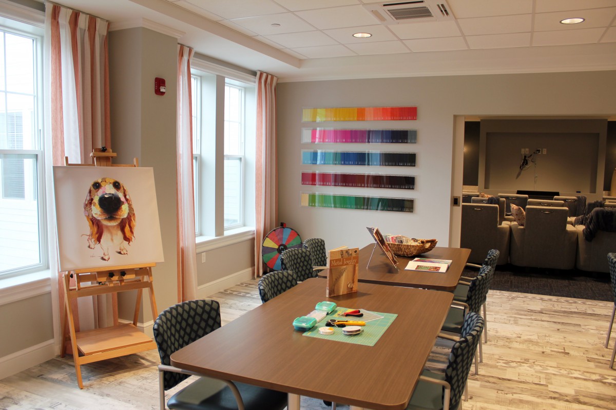 A well-lit art room with a large table, chairs, and painting supplies, featuring colorful artwork on the wall.