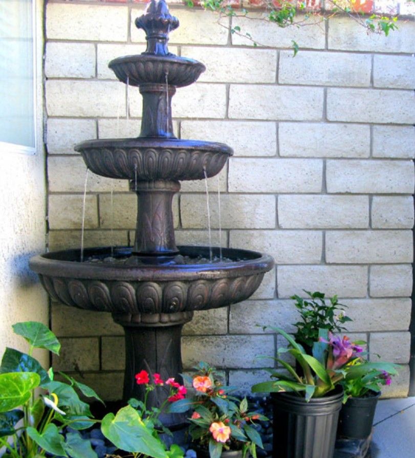 A three-tier water fountain surrounded by colorful potted plants, set against a brick wall.