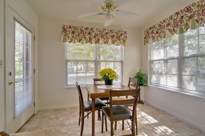 A bright dining area with natural light, featuring a wooden table and chairs surrounded by large windows with floral curtains.