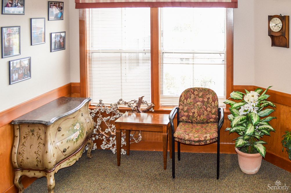 A cozy interior corner featuring a window with sheer curtains, a patterned chair, a wooden table, and decorative items.