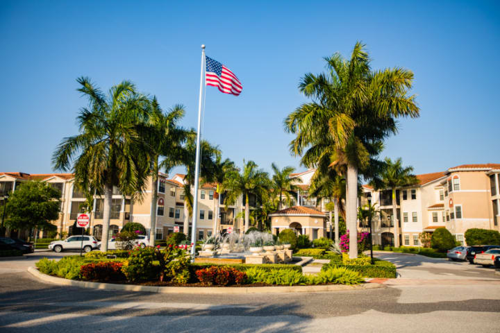 A scenic view of an apartment complex with palm trees and an American flag on a sunny day.