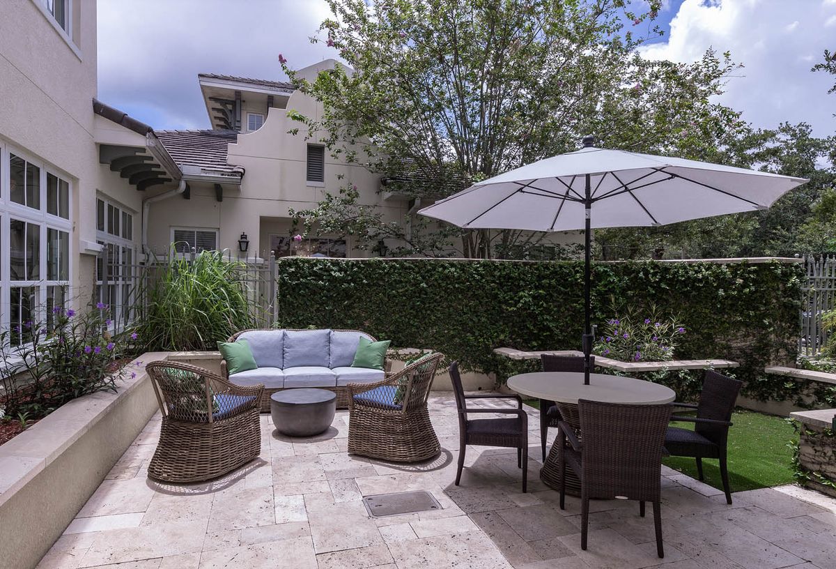 A cozy patio with a seating area, table, and umbrella next to a green hedge in a backyard.
