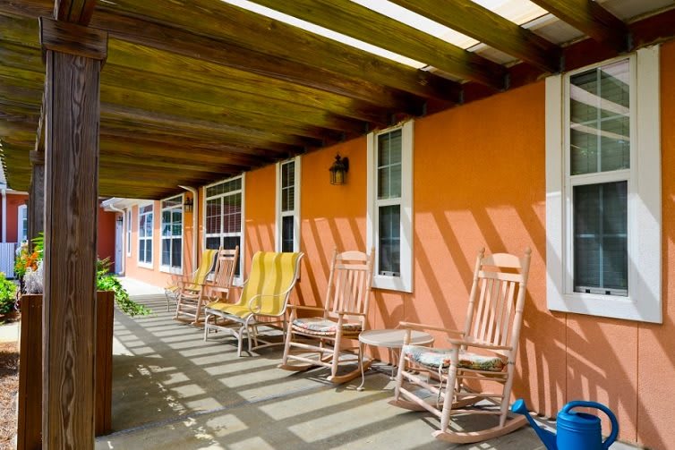 A row of wooden rocking chairs on a porch with an orange wall, basking in sunlight and shade.