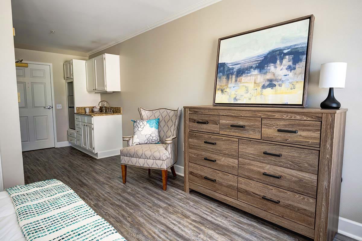 A modern bedroom featuring a wooden dresser, patterned armchair, and abstract artwork on the wall.