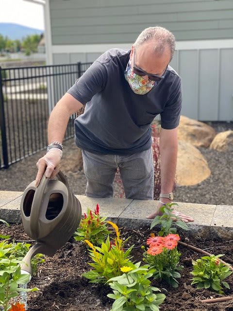 A person wearing a mask waters plants in a garden.