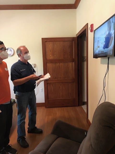 Two people wearing face masks are discussing in front of a television screen indoors.