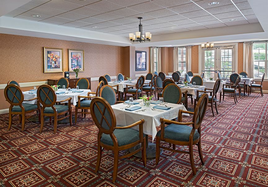 A spacious dining room with elegantly set tables and chairs, featuring a patterned carpet and decorative chandeliers.