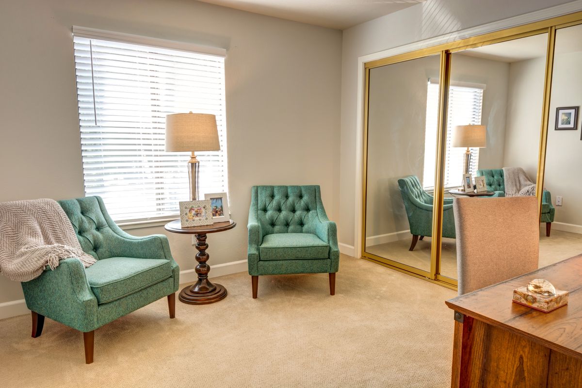 A cozy living room with two teal chairs, a round wooden table, and a large mirror.