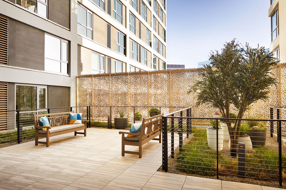 A modern outdoor patio area with wooden benches, greenery, and urban architecture.