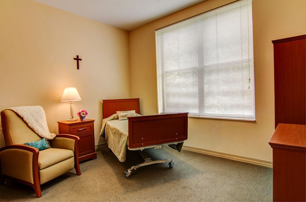 A simple room featuring a hospital bed next to a side table with a lamp, a comfortable chair, and a window with blinds. A cross is mounted on the wall.