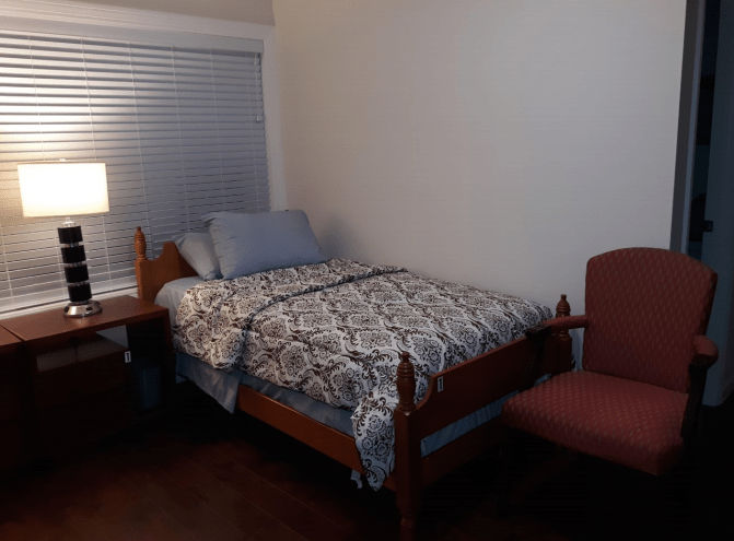 A cozy bedroom with a single bed, nightstand with a lamp, and an upholstered chair.
