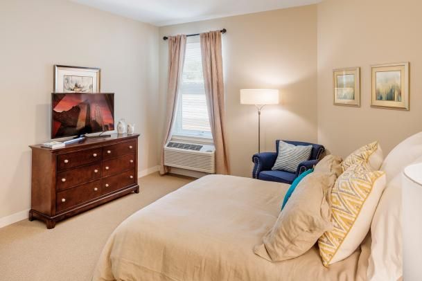 A cozy bedroom with a neatly made bed, a wooden dresser with a TV on top, and a comfortable armchair by the window.