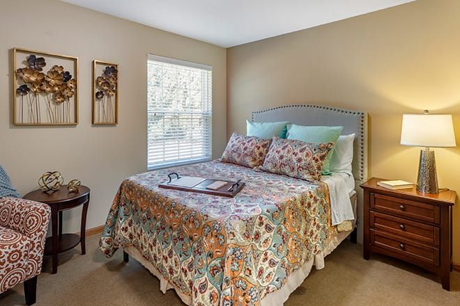 A cozy bedroom with a patterned bedspread and soft pillows, featuring a wooden nightstand and a window letting in natural light.