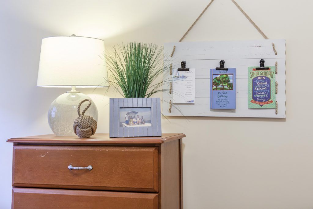 A cozy interior scene featuring a lamp, picture frame, and plants on a dresser with decorative wall art.