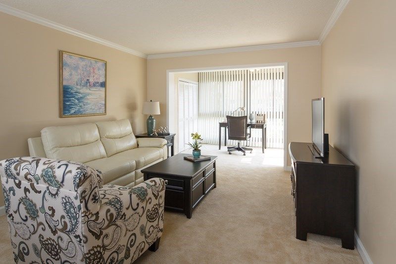 A cozy living room with a sofa, patterned armchair, and coffee table, featuring a desk near a window with blinds.