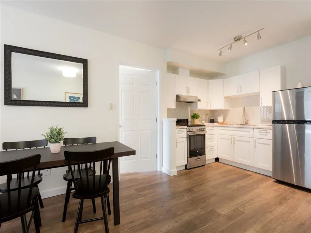 A modern minimalist kitchen with white cabinets and a dining area featuring wood flooring.