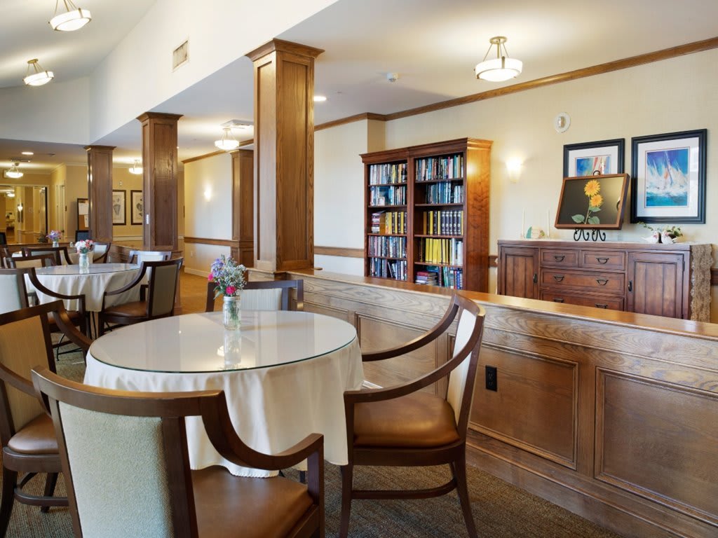 A cozy dining area with round tables, high-backed chairs, and a bookshelf against wood-paneled walls.