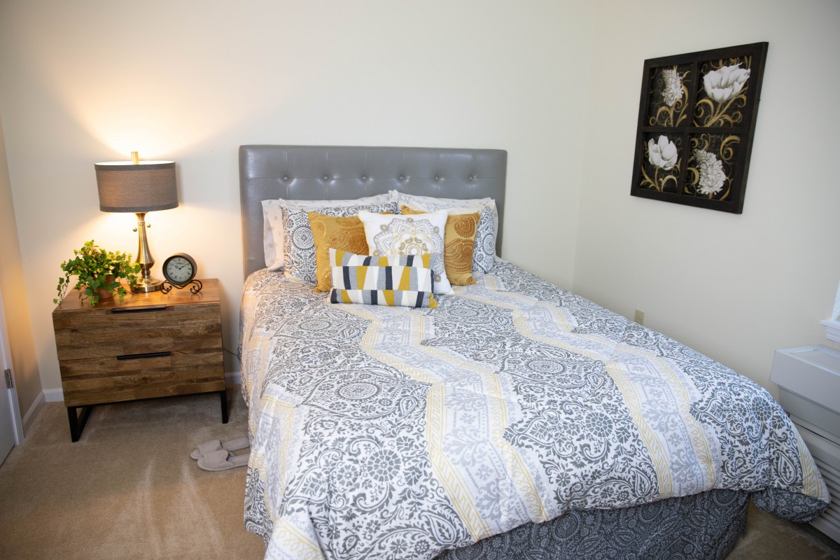 A cozy bedroom with a grey upholstered bed, patterned bedding, decorative pillows, and a wooden nightstand with a lamp.