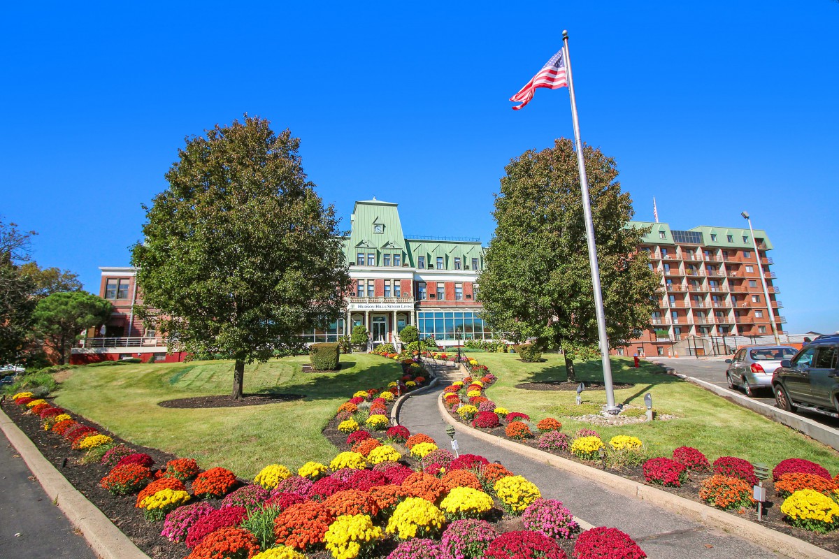 A beautifully landscaped garden with colorful flowers leading to a large building, accompanied by a flagpole and trees under a clear blue sky.